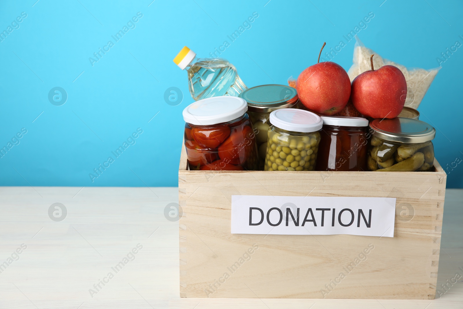 Photo of Different food products for donation in wooden crate on table against light blue background, space for text