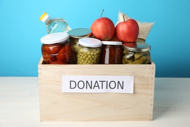 Photo of Different food products for donation in wooden crate on table against light blue background