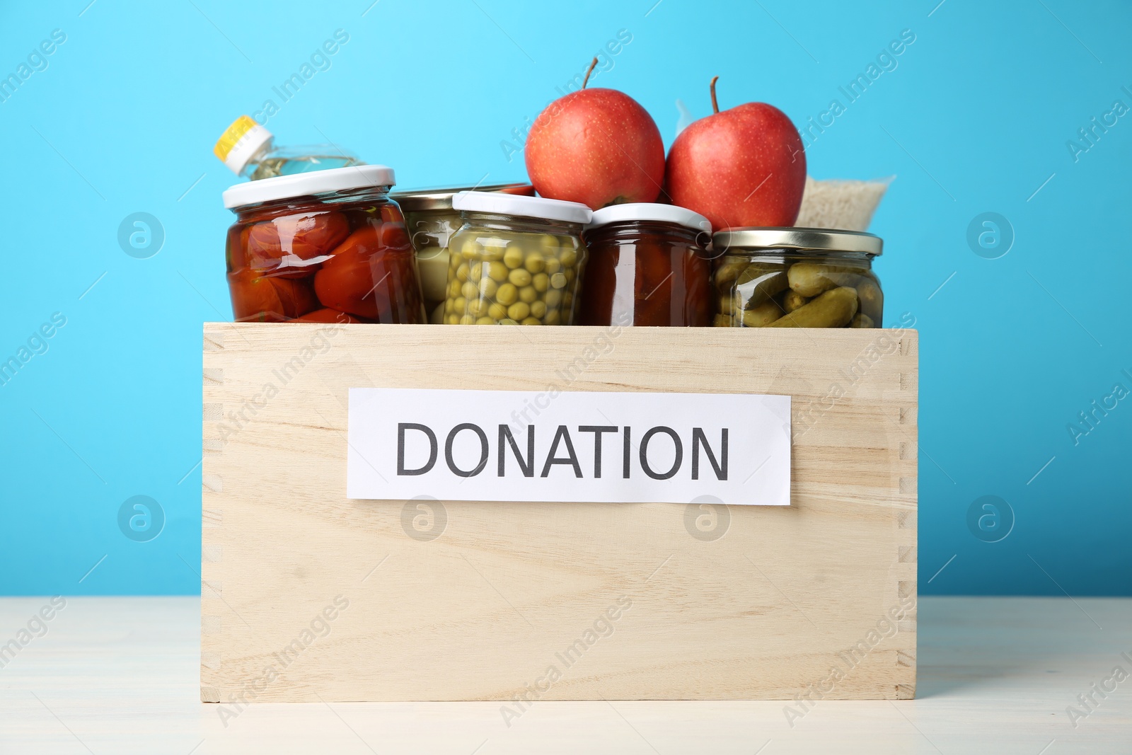 Photo of Different food products for donation in wooden crate on table against light blue background