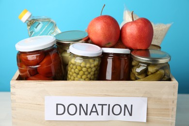 Photo of Different food products for donation in wooden crate on table against light blue background, closeup