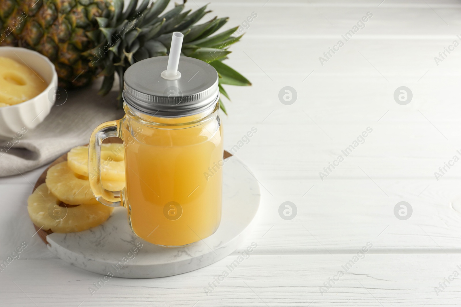 Photo of Tasty pineapple juice in mason jar and slices of fresh fruit on white wooden table, closeup. Space for text