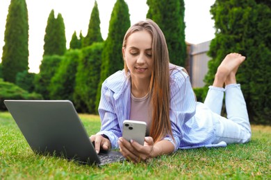 Woman with smartphone using laptop on green lawn in park