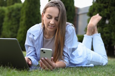 Woman with smartphone using laptop on green lawn in park