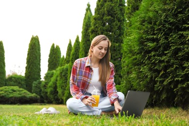 Woman with juice using laptop on green lawn in park