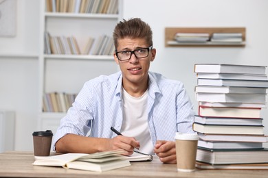 Photo of Tired student before exam with stack of books and coffee at table indoors