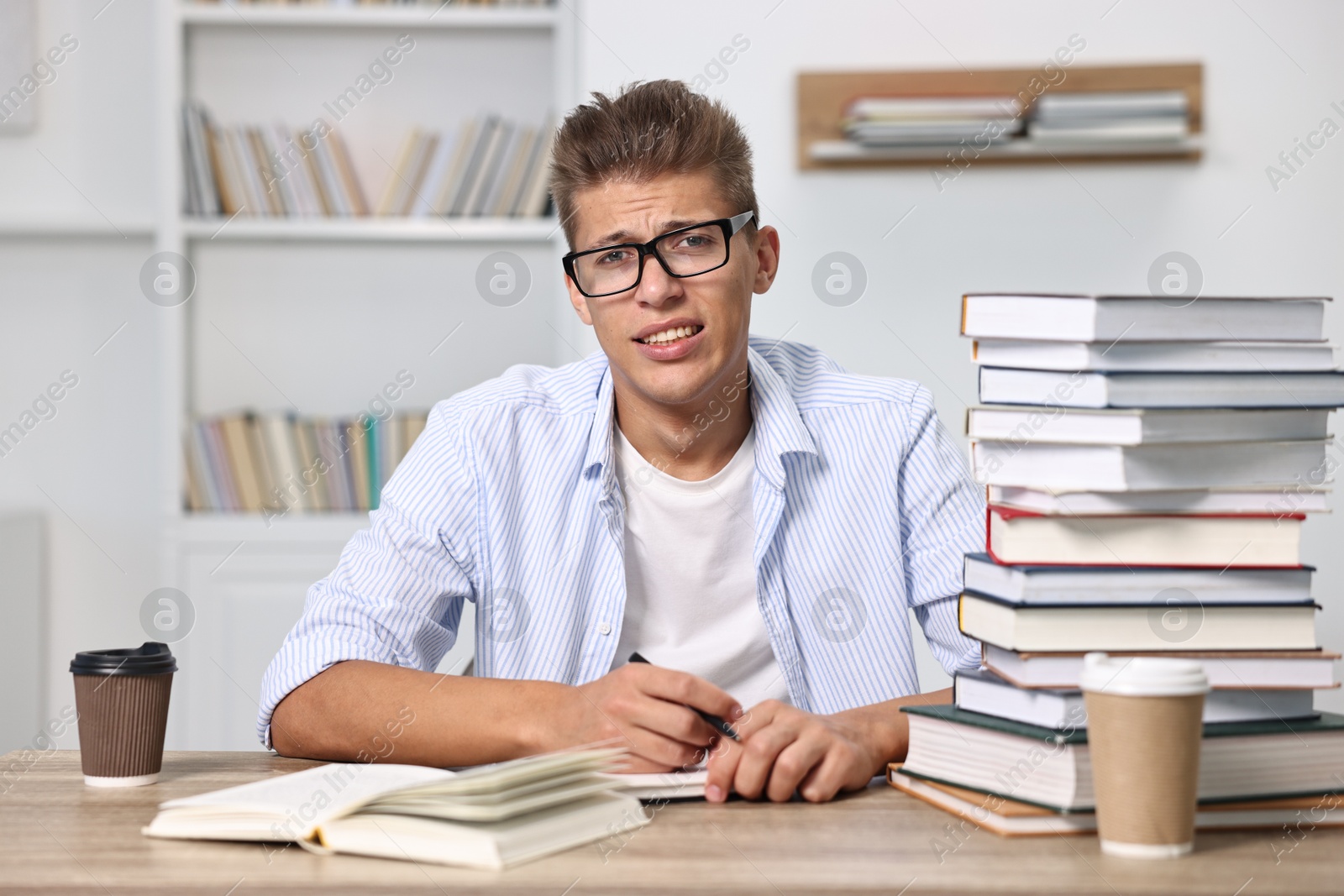 Photo of Tired student before exam with stack of books and coffee at table indoors