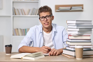 Tired student before exam with stack of books and coffee at table indoors