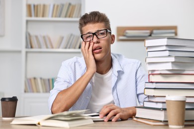 Photo of Tired student before exam with stack of books and coffee at table indoors