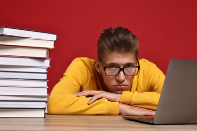 Photo of Tired student before exam with stack of books and laptop at wooden table against red background