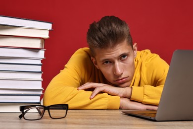 Photo of Tired student before exam with stack of books and laptop at wooden table against red background