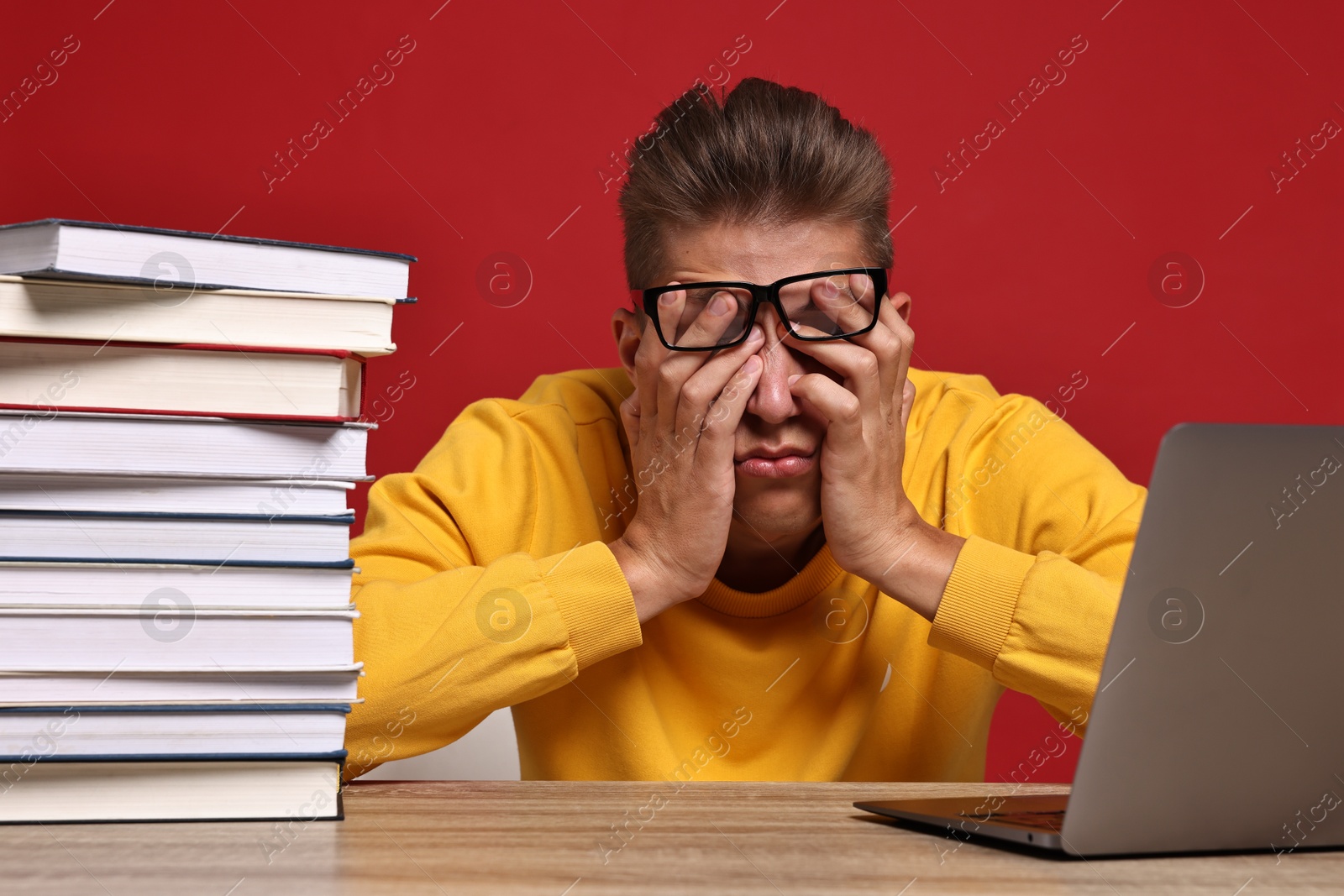 Photo of Tired student before exam with stack of books and laptop at wooden table against red background