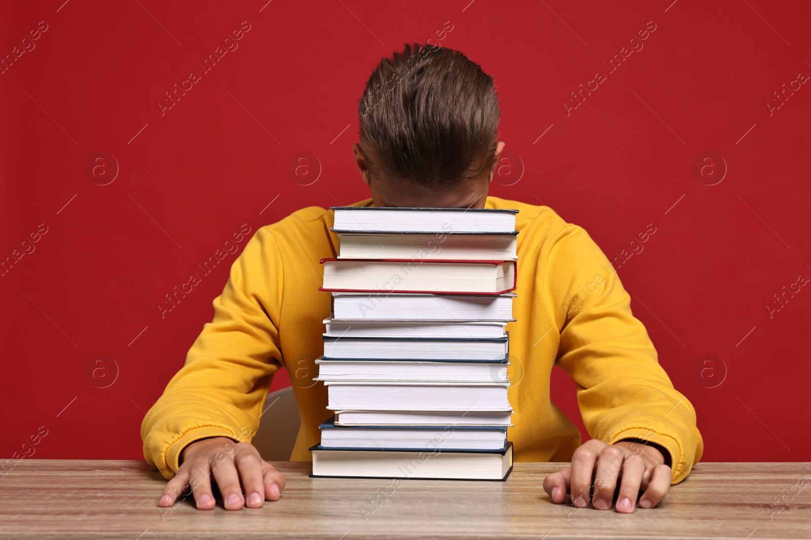 Photo of Tired student before exam with stack of books at wooden table against red background