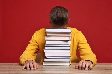 Tired student before exam with stack of books at wooden table against red background