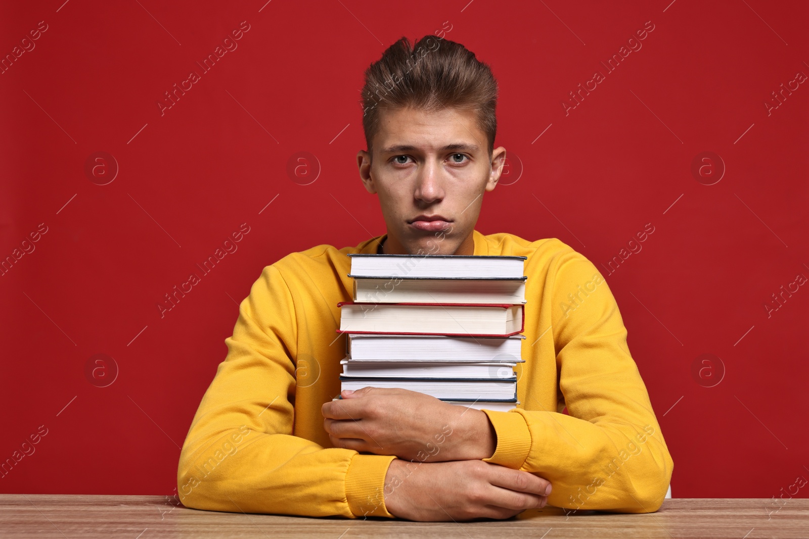 Photo of Tired student before exam with stack of books at wooden table against red background