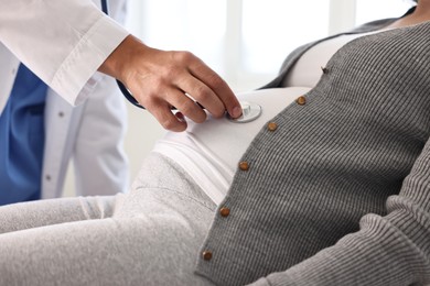 Pregnancy checkup. Doctor with stethoscope listening baby's heartbeat in patient's tummy indoors, closeup