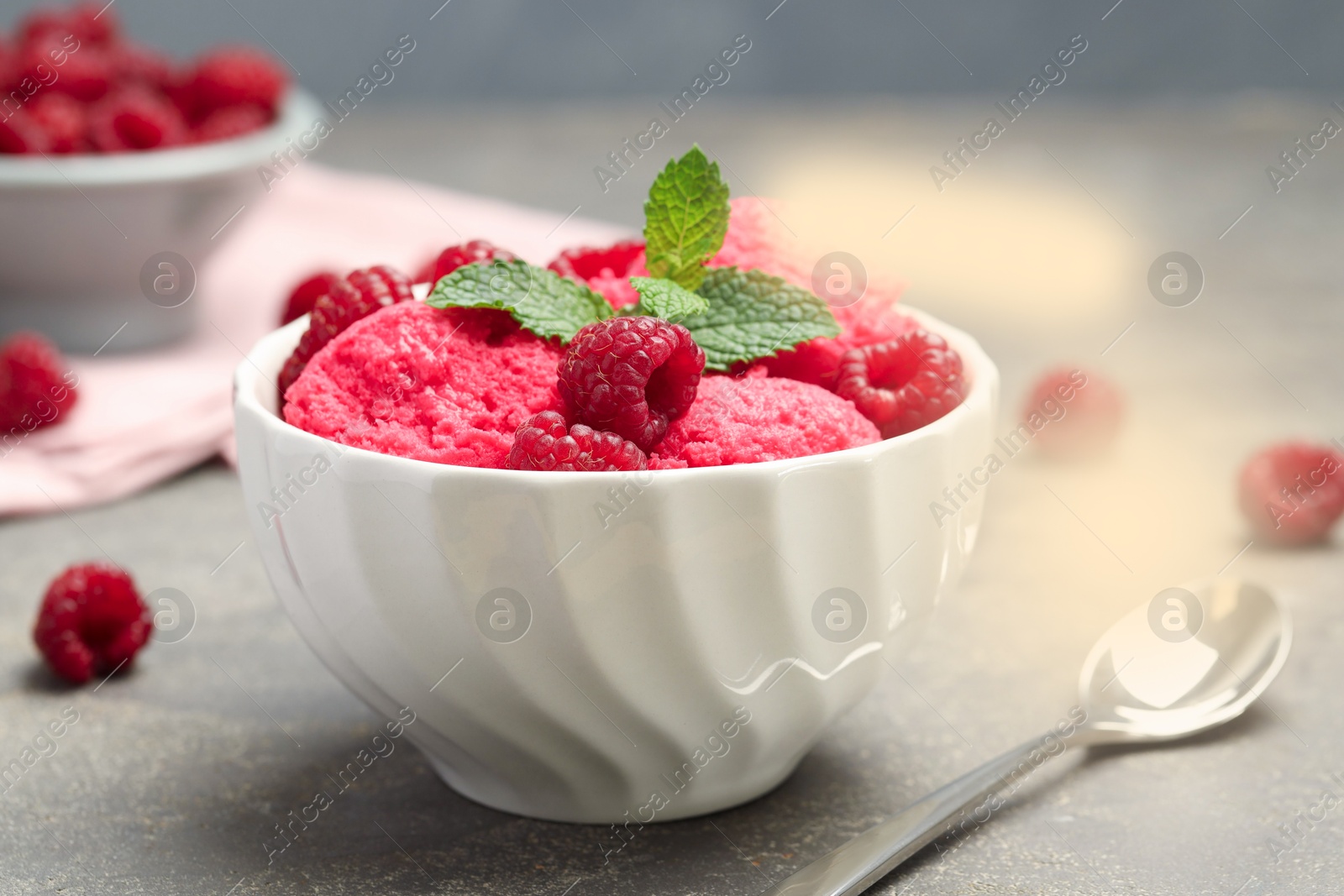 Photo of Delicious raspberry sorbet with fresh berries in bowl and spoon on gray textured table, closeup