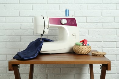 Sewing machine, blue fabric and spools of threads on wooden table near white brick wall