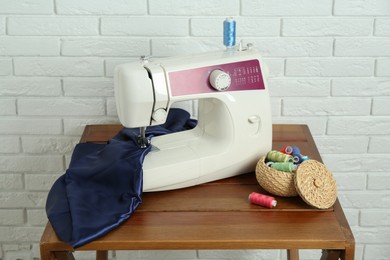 Photo of Sewing machine, blue fabric and spools of threads on wooden table near white brick wall