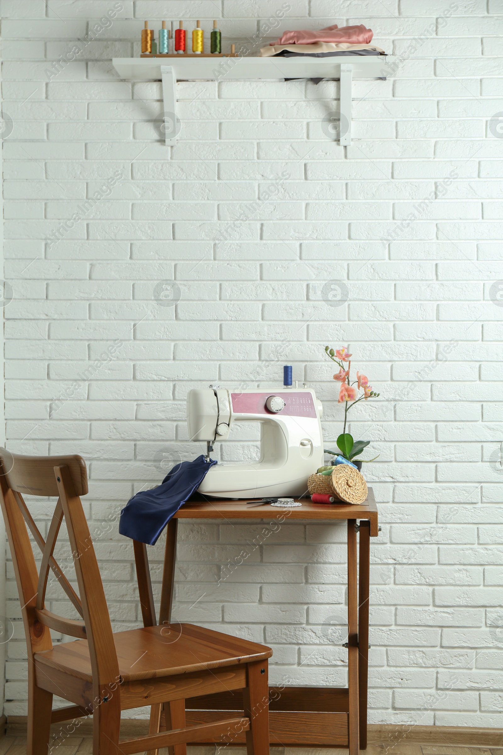 Photo of Sewing machine, blue fabric and craft accessories on wooden table near white brick wall