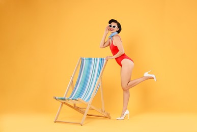 Pin-up woman in swimsuit near folding chair on orange background