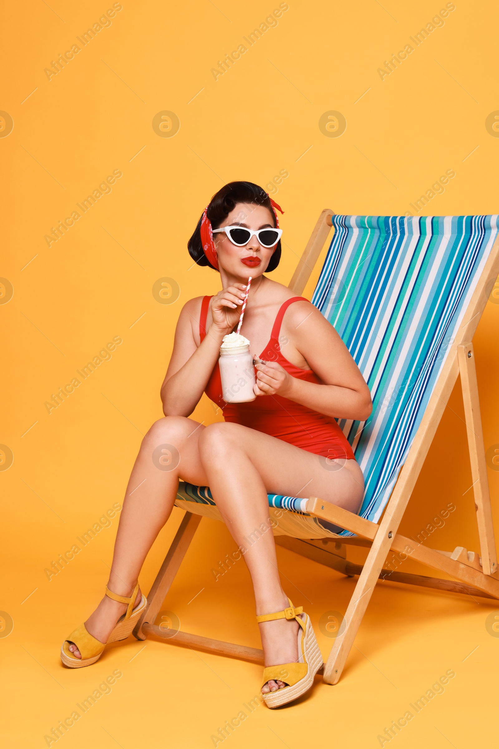 Photo of Pin-up woman in swimsuit with milk shake on folding chair against orange background