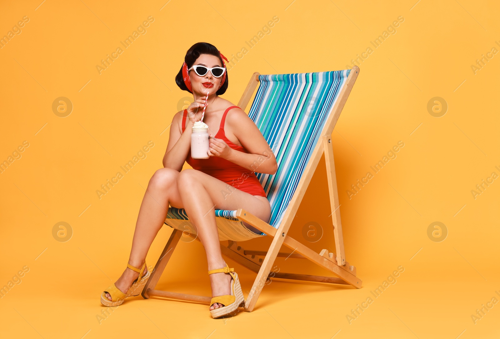 Photo of Pin-up woman in swimsuit with milk shake on folding chair against orange background