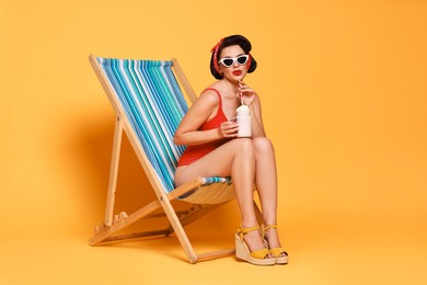 Photo of Pin-up woman in swimsuit with milk shake on folding chair against orange background