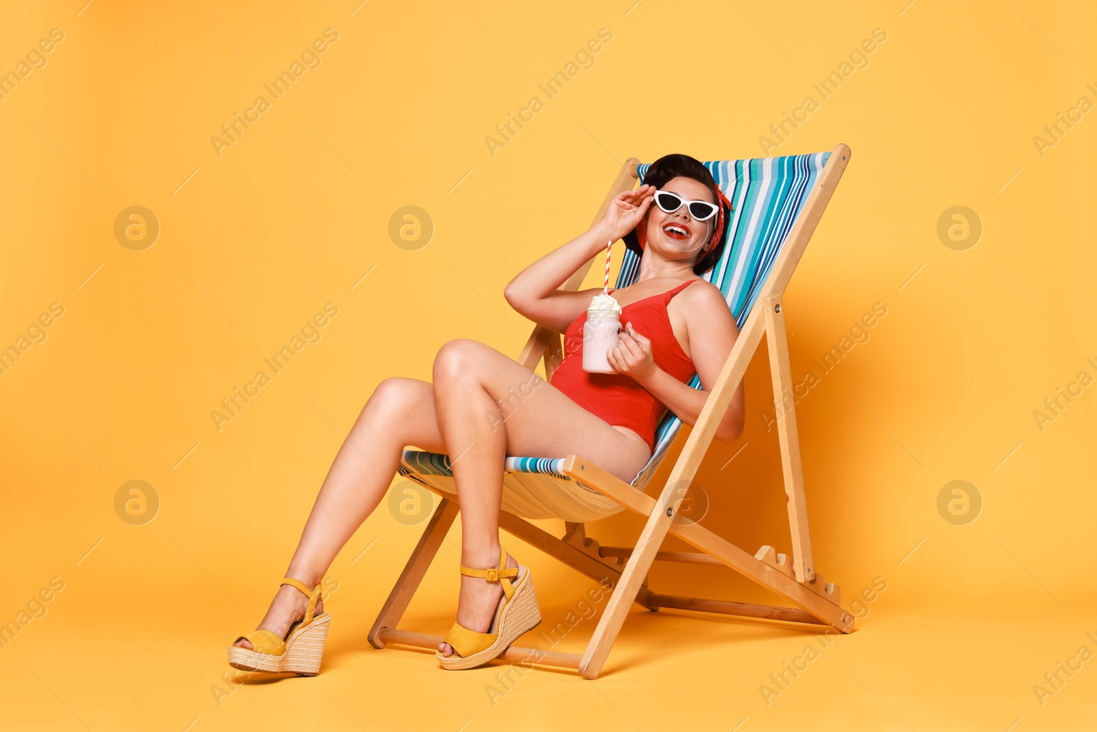 Photo of Pin-up woman in swimsuit with milk shake on folding chair against orange background