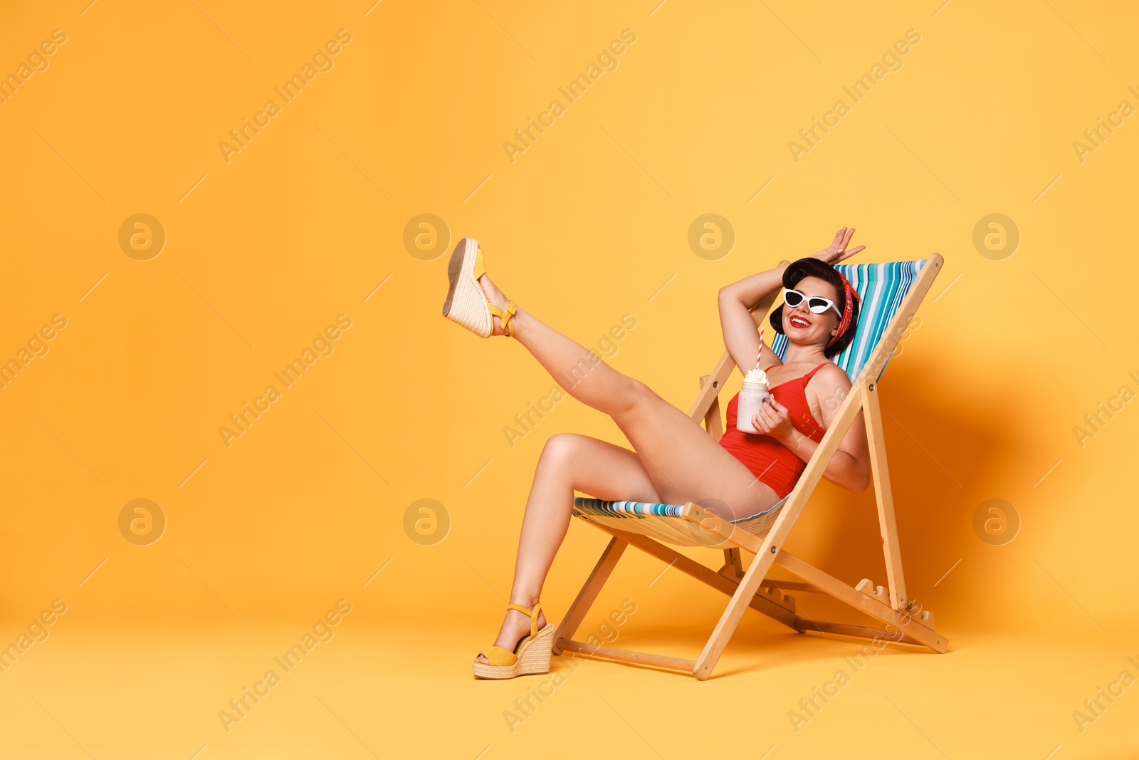 Photo of Pin-up woman in swimsuit with milk shake on folding chair against orange background, space for text