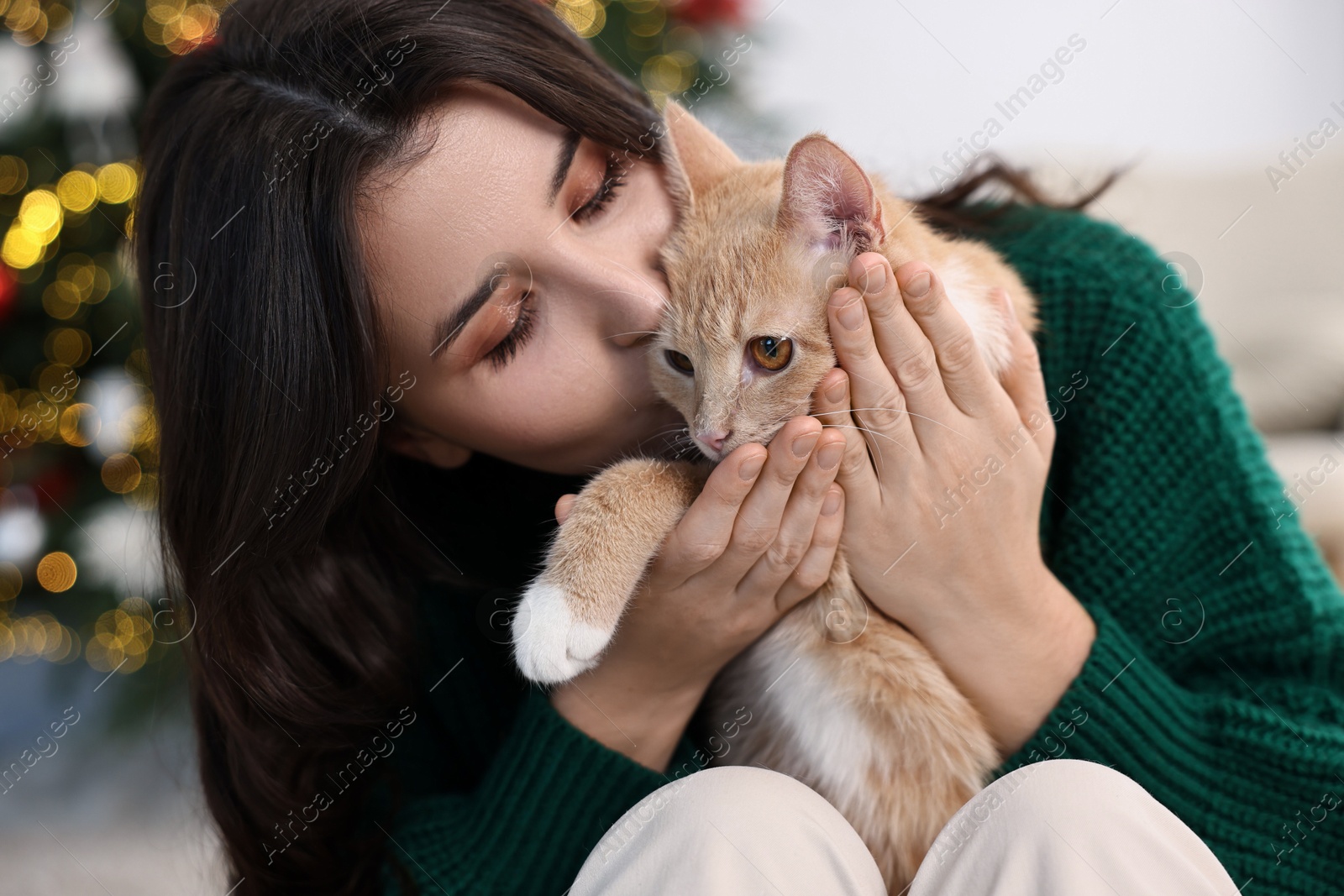 Photo of Woman kissing her cute ginger cat in room decorated for Christmas