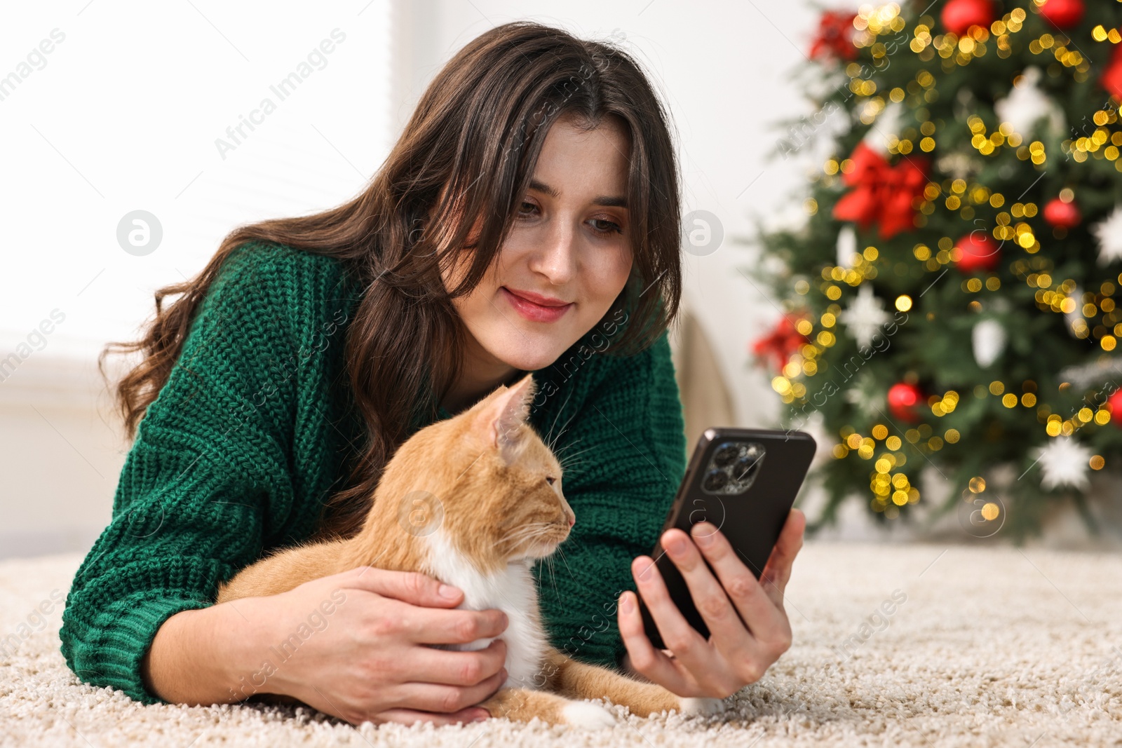 Photo of Woman using smartphone and her cute ginger cat lying on rug in room decorated for Christmas