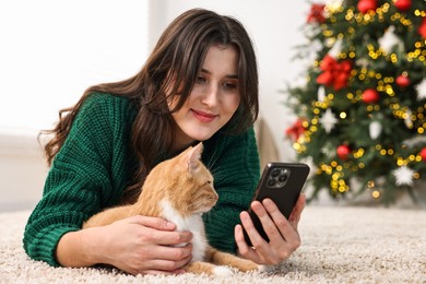 Photo of Woman using smartphone and her cute ginger cat lying on rug in room decorated for Christmas