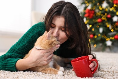 Woman kissing her cute ginger cat on rug in room decorated for Christmas