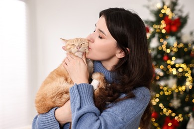 Photo of Woman kissing her cute ginger cat in room decorated for Christmas