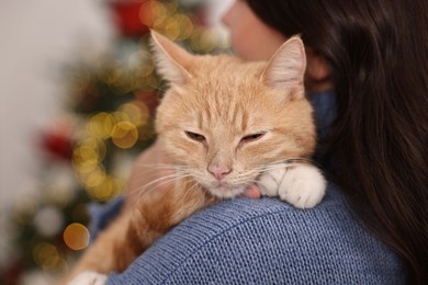 Photo of Woman with cute ginger cat against blurred Christmas lights indoors, closeup