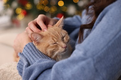 Photo of Woman with cute ginger cat against blurred Christmas lights indoors, closeup