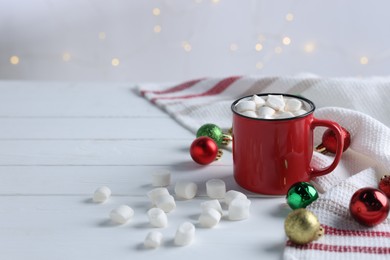 Photo of Yummy cocoa with marshmallows in red cup and Christmas balls on white wooden table, space for text