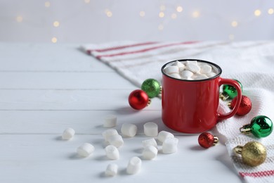 Photo of Yummy cocoa with marshmallows in red cup and Christmas balls on white wooden table, space for text