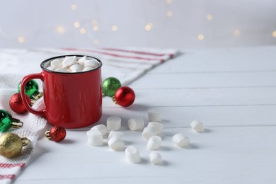 Photo of Yummy cocoa with marshmallows in red cup and Christmas balls on white wooden table, space for text