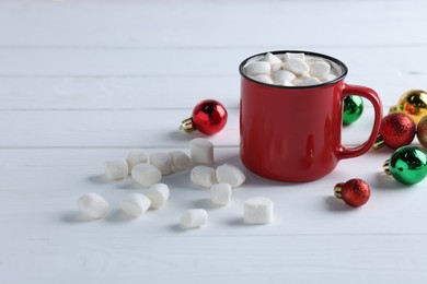 Photo of Yummy cocoa with marshmallows in red cup and Christmas balls on white wooden table, space for text