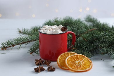 Photo of Yummy cocoa with marshmallows in red cup, anise stars, dried orange slices and fir tree branches on white wooden table