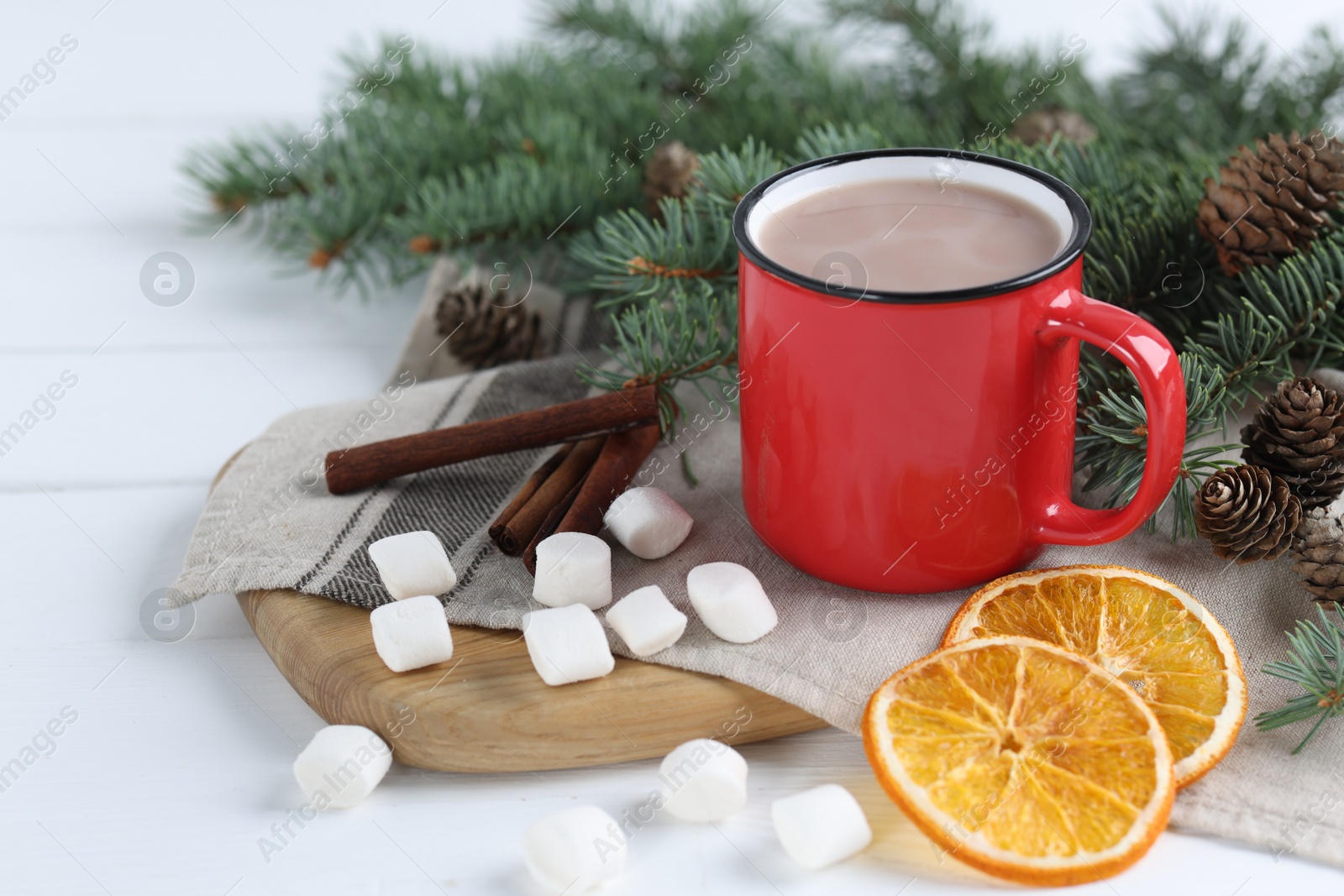 Photo of Yummy cocoa in red cup, marshmallows, cinnamon sticks, dried orange slices and fir tree branches on white wooden table. Space for text