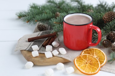 Photo of Yummy cocoa in red cup, marshmallows, cinnamon sticks, dried orange slices and fir tree branches on white wooden table. Space for text