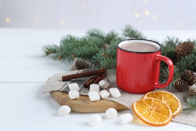 Photo of Yummy cocoa in red cup, marshmallows, cinnamon sticks, dried orange slices and fir tree branches on white wooden table. Space for text