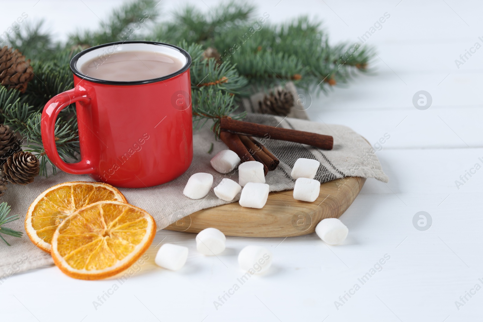 Photo of Yummy cocoa in red cup, marshmallows, cinnamon sticks, dried orange slices and fir tree branches on white wooden table. Space for text