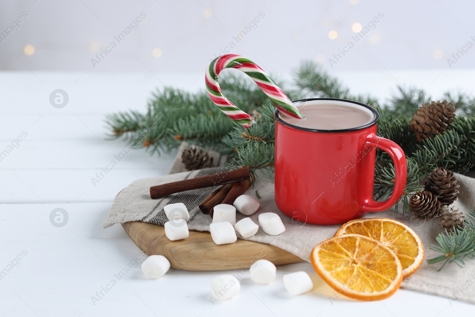 Photo of Yummy cocoa and candy cane in red cup, marshmallows, cinnamon sticks, dried orange slices and fir tree branches on white wooden table. Space for text