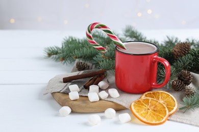 Photo of Yummy cocoa and candy cane in red cup, marshmallows, cinnamon sticks, dried orange slices and fir tree branches on white wooden table. Space for text