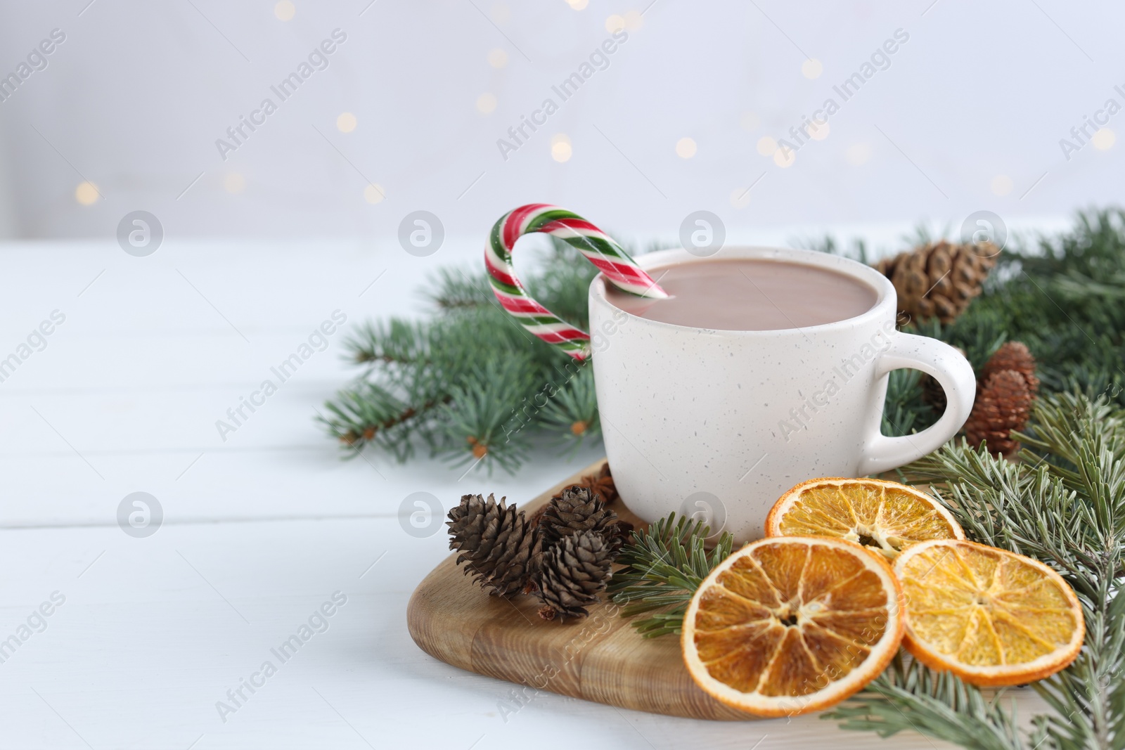 Photo of Yummy cocoa and candy cane in cup, dried orange slices and fir tree branches on white wooden table. Space for text