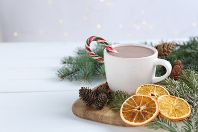 Photo of Yummy cocoa and candy cane in cup, dried orange slices and fir tree branches on white wooden table. Space for text