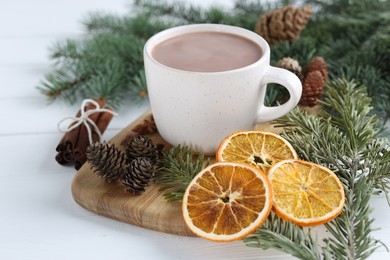 Photo of Yummy cocoa in cup, dried orange slices, cinnamon sticks and fir tree branches on white wooden table, closeup